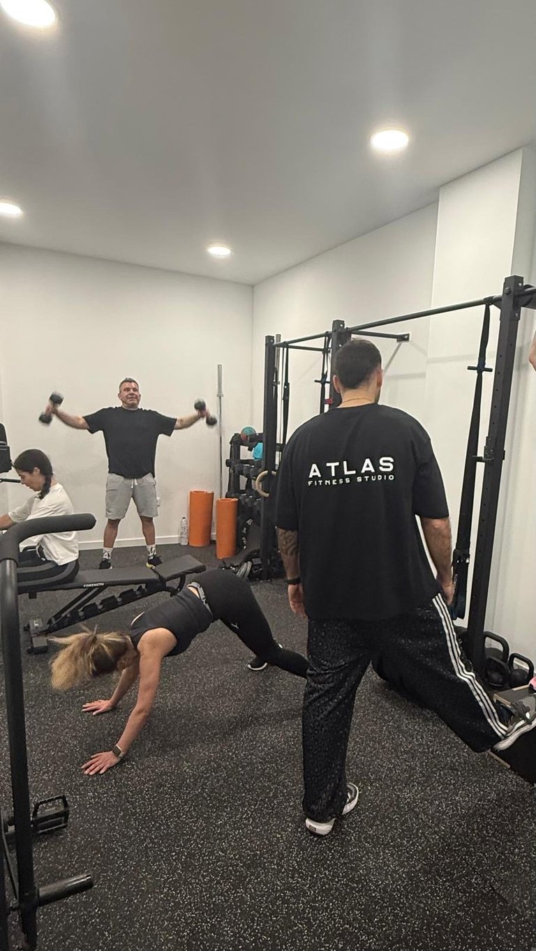 People exercising in a modern gym. A woman does a plank while a man in an Atlas shirt watches. Another man lifts dumbbells in the background.