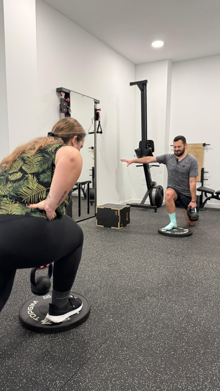 A trainer instructing a woman performing a balance board exercise in a modern fitness gym studio with mirrored walls